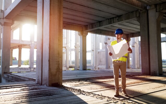 Ingénieur civil avec casque et gilet de sécurité vérifie les plans du chantier dans un gros œuvre à plusieurs étages au coucher du soleil