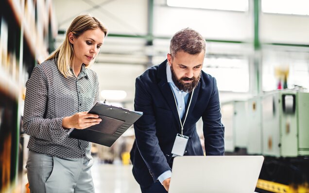 Deux spécialistes en discussion dans un hall industriel moderne avec un ordinateur portable et une planchette à pince
